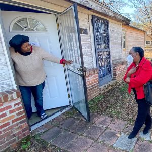 Woman leans out of the doorway of her older home and speaks to a woman standing on the home's walkway