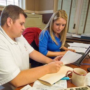 A man and woman work together while sitting at a desk covered in documents