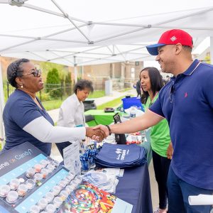 Two people under outdoor tent shake hands across an event table