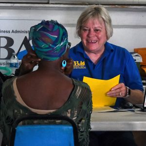 Two women sit across from each other at a table at an event while one holds a piece of paper