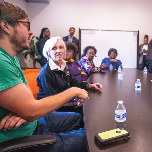 Young man speaking at a boardroom table with an audience in the background