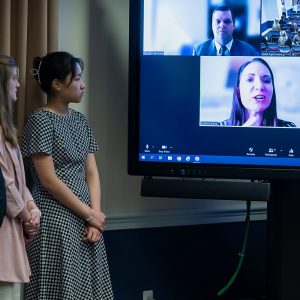 Young woman standing next to TV screen with faces on a video conference