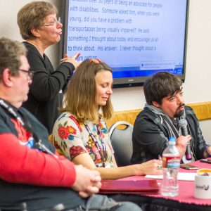 Panelists at a meeting with a sign language interpreter and a typed transcript on a screen behind them