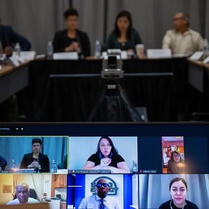 An in-person meeting with a laptop in the foreground showing people on a video conference